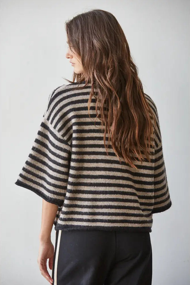 A woman with long brown hair is shown from the back wearing a loose, short-sleeved knit top with horizontal black and beige stripes, paired with black pants with white side stripes, against a light gray studio background.