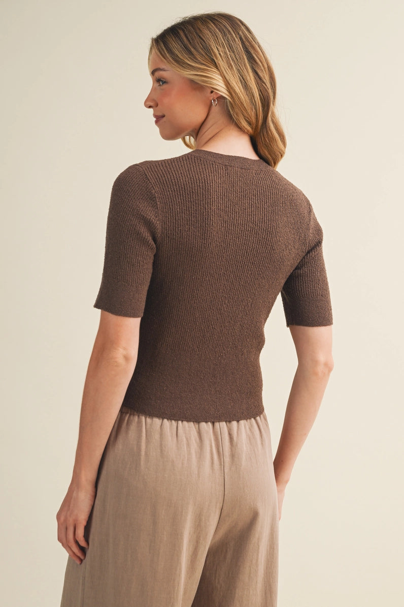 A woman shown from the back wearing a fitted short-sleeve ribbed knit top in a medium brown color, paired with light beige wide-leg trousers, against a plain beige studio background.