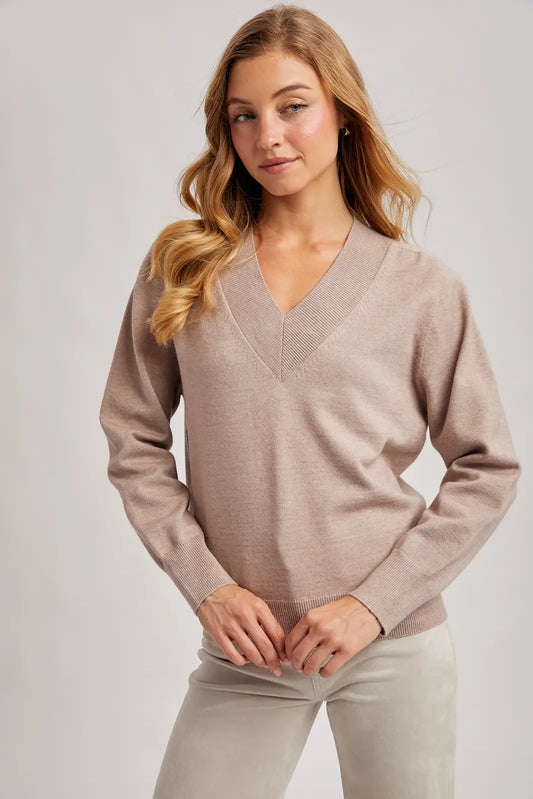A woman wearing a light taupe knit V-neck sweater and light beige pants, standing against a neutral light gray studio background.