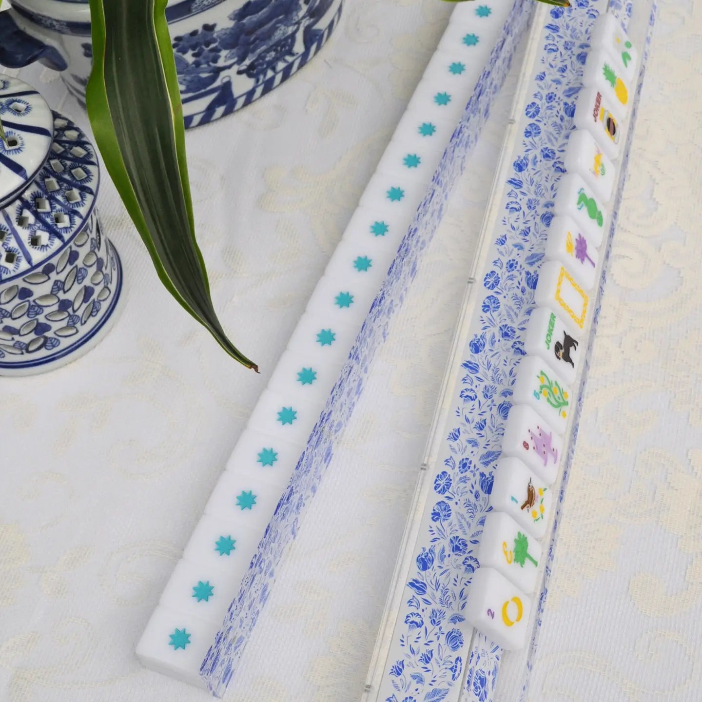 A set of white mahjong tiles with blue floral patterns and turquoise accents, displayed in a clear case on a white patterned tablecloth with decorative blue-and-white ceramic vessels and a green leaf.