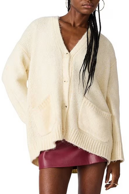 A woman wearing an oversized cream-colored fuzzy knit cardigan with a V-neck, button front, and large front pockets, paired with a dark red skirt, against a plain white background.