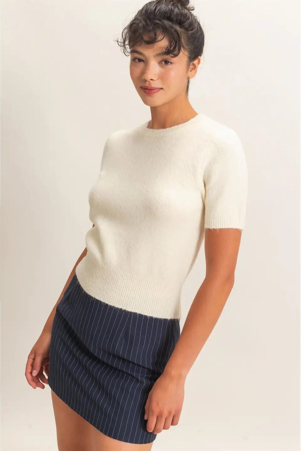 A woman modeling a cream short-sleeve knit top paired with a navy blue and white pinstriped mini skirt, set against a plain light beige studio background.