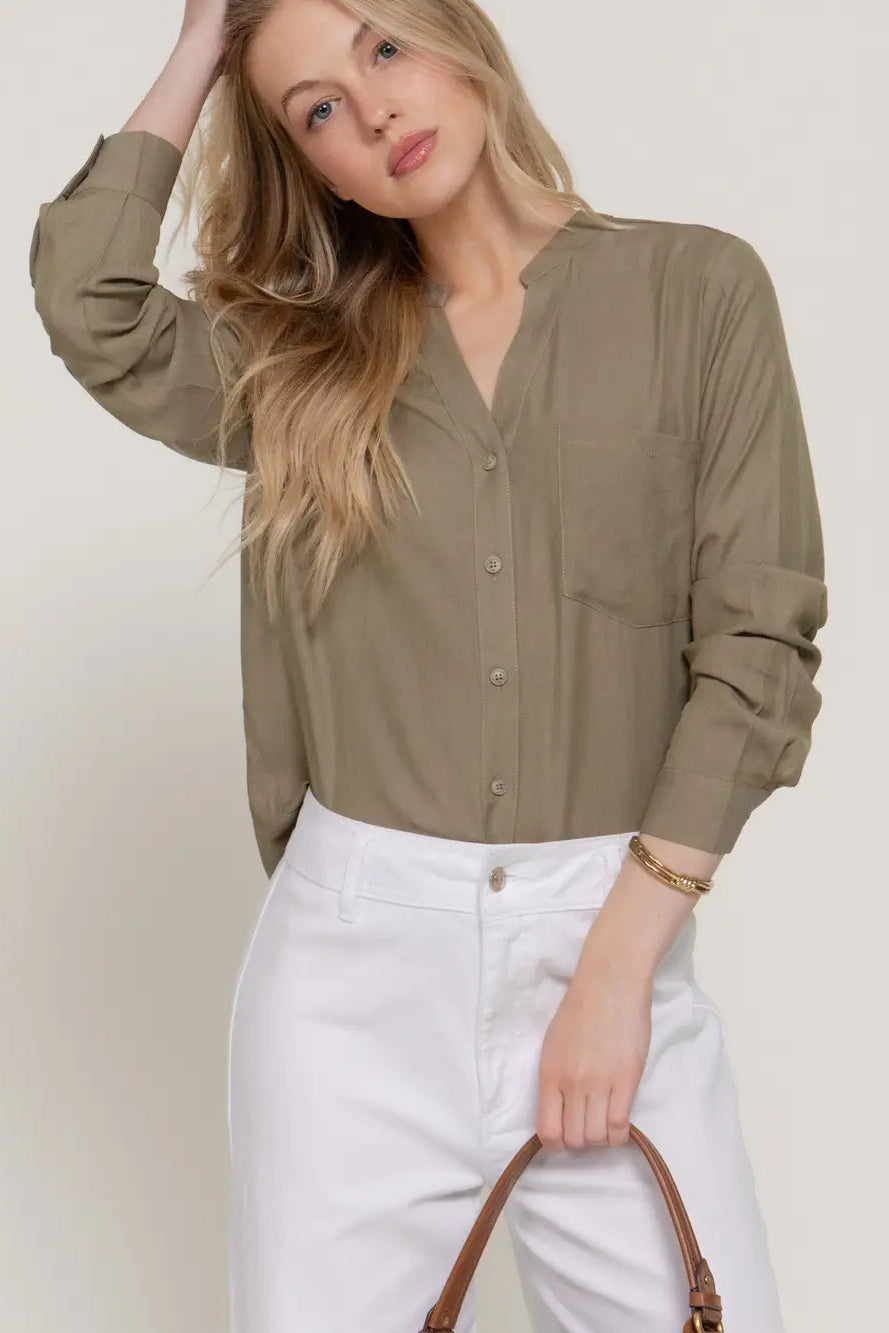 A woman wearing a long-sleeve button-front blouse in a muted olive khaki color, paired with white pants, holding a brown leather handbag against a plain light beige studio background.