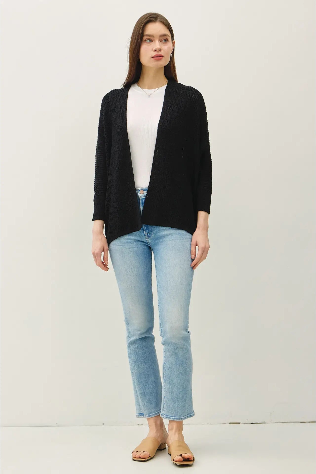 A woman modeling a black open-front knit cardigan over a white top, paired with light-wash blue jeans and tan sandals, against a plain light gray studio background.