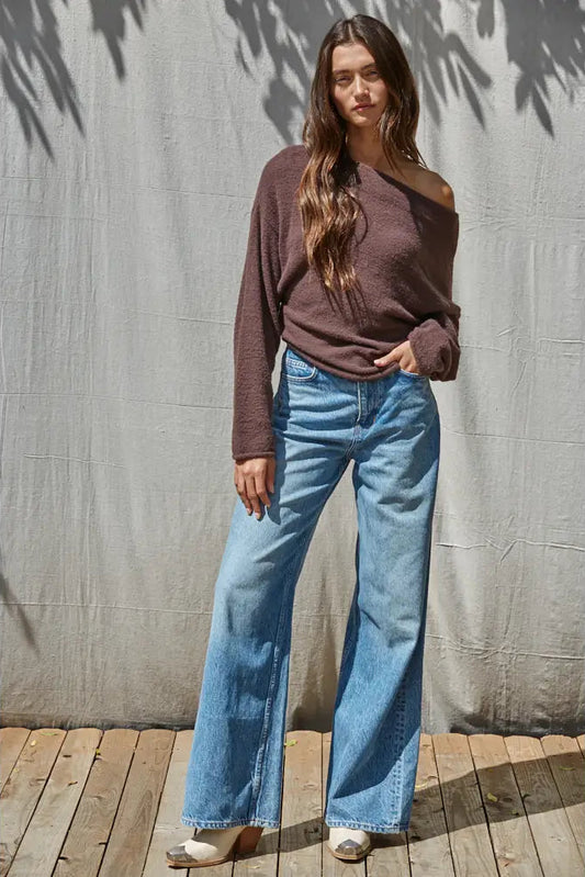 A woman stands outdoors against a gray fabric backdrop on a wooden deck, wearing a brown off-the-shoulder knit sweater and light blue wide-leg jeans.