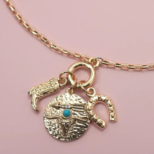 Gold necklace with longhorn bull, turquoise stone, and horseshoe charm on a pink background