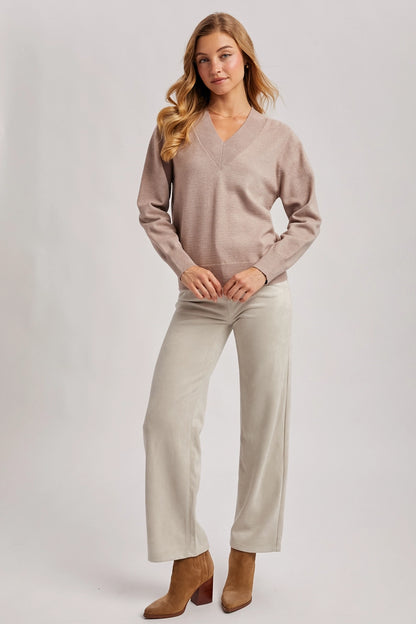 A woman modeling a light taupe long-sleeve V-neck knit sweater and light beige wide-leg trousers, paired with brown ankle boots, against a light gray studio background.
