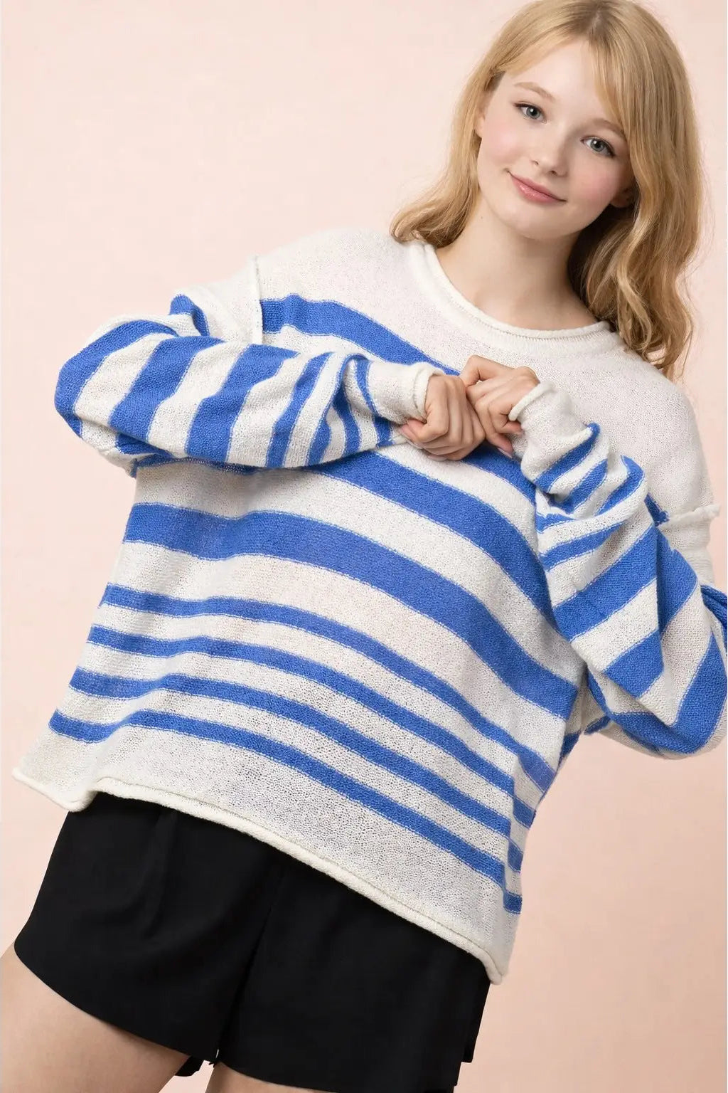 A young woman wearing a blue and white striped knit sweater with black shorts, posing against a solid pale pink background.