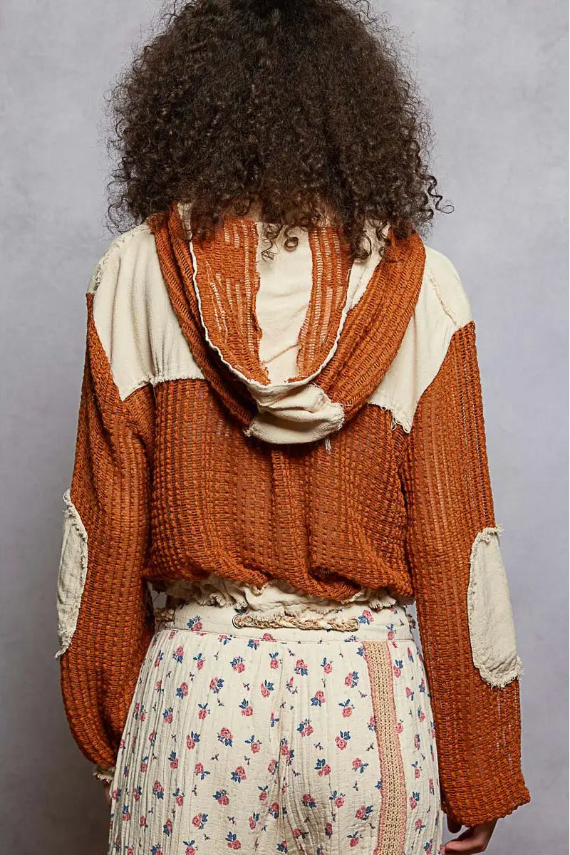 Person wearing a brown knitted sweater over a cream shirt with a floral skirt, standing against a gray background.
