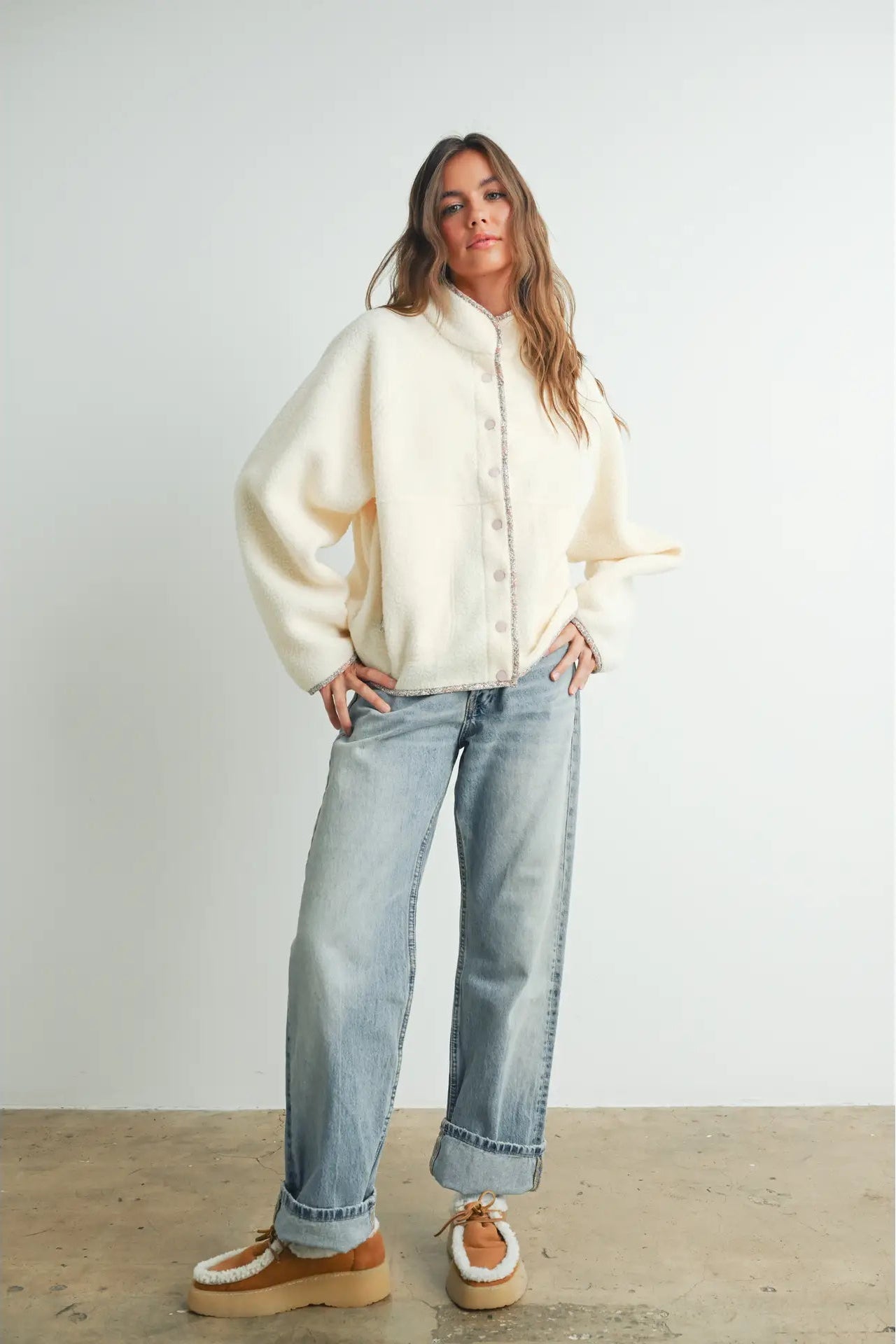 A woman modeling a cream-colored, oversized fuzzy button-up cardigan with wide sleeves, paired with light-wash blue jeans and brown platform shoes, standing against a plain light gray wall.