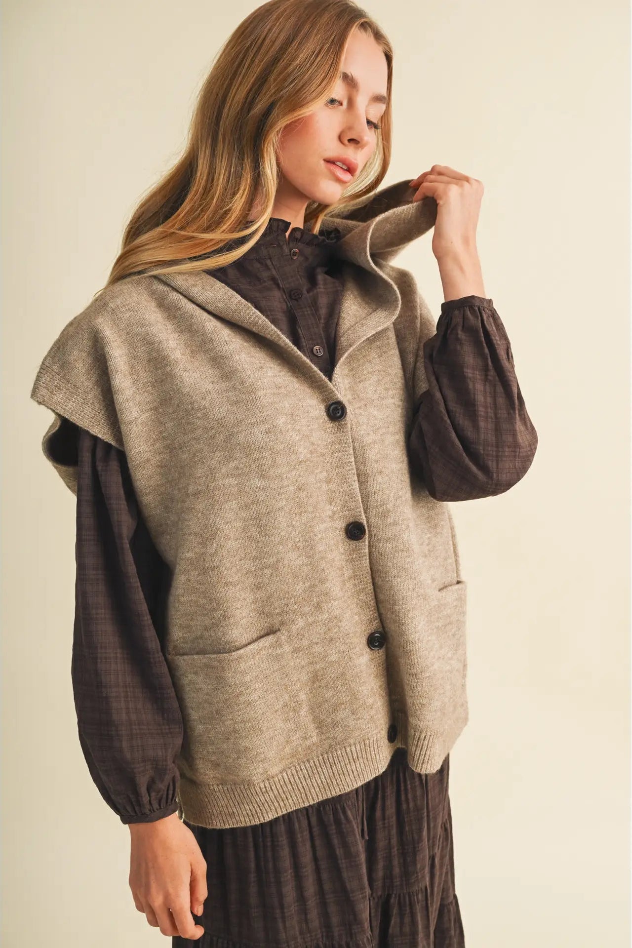 A woman wearing a beige sleeveless knit cardigan vest over a dark brown long-sleeve shirt and dark brown skirt, posing against a solid light beige background.