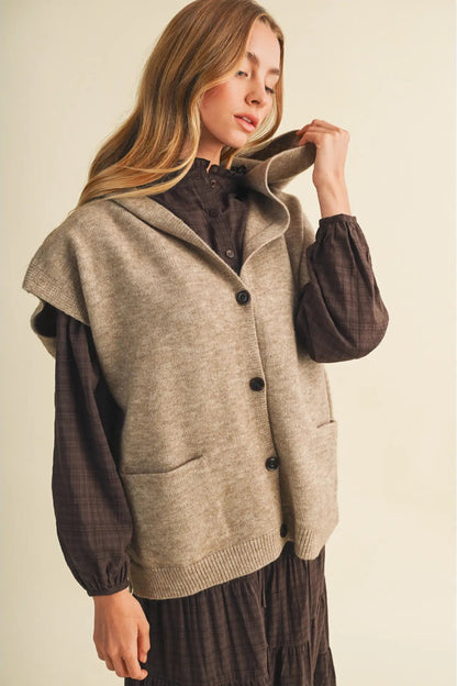 A woman wearing a beige sleeveless knit cardigan vest over a dark brown long-sleeve shirt and dark brown skirt, posing against a solid light beige background.