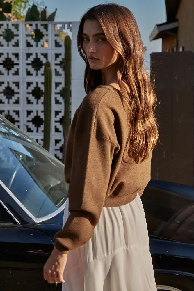 A woman wearing a brown knit crewneck sweater and a light cream skirt, standing outdoors next to a black car with a decorative wall in the background.