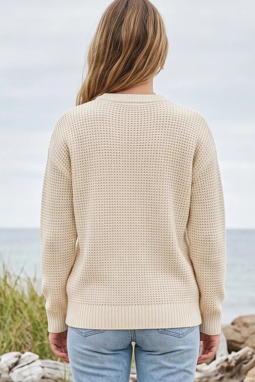 A woman viewed from behind wearing a cream-colored waffle-knit sweater and light blue jeans, standing outdoors near the ocean with rocks and grass in the background.