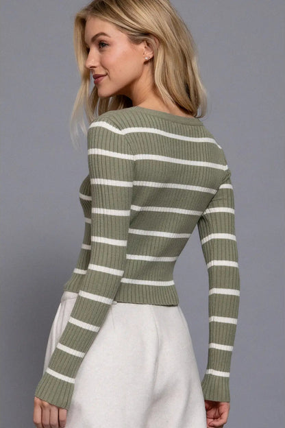 A woman wearing a ribbed long-sleeve sweater with horizontal olive green and white stripes, paired with a light gray skirt, shown from a three-quarter back angle against a solid gray studio background.
