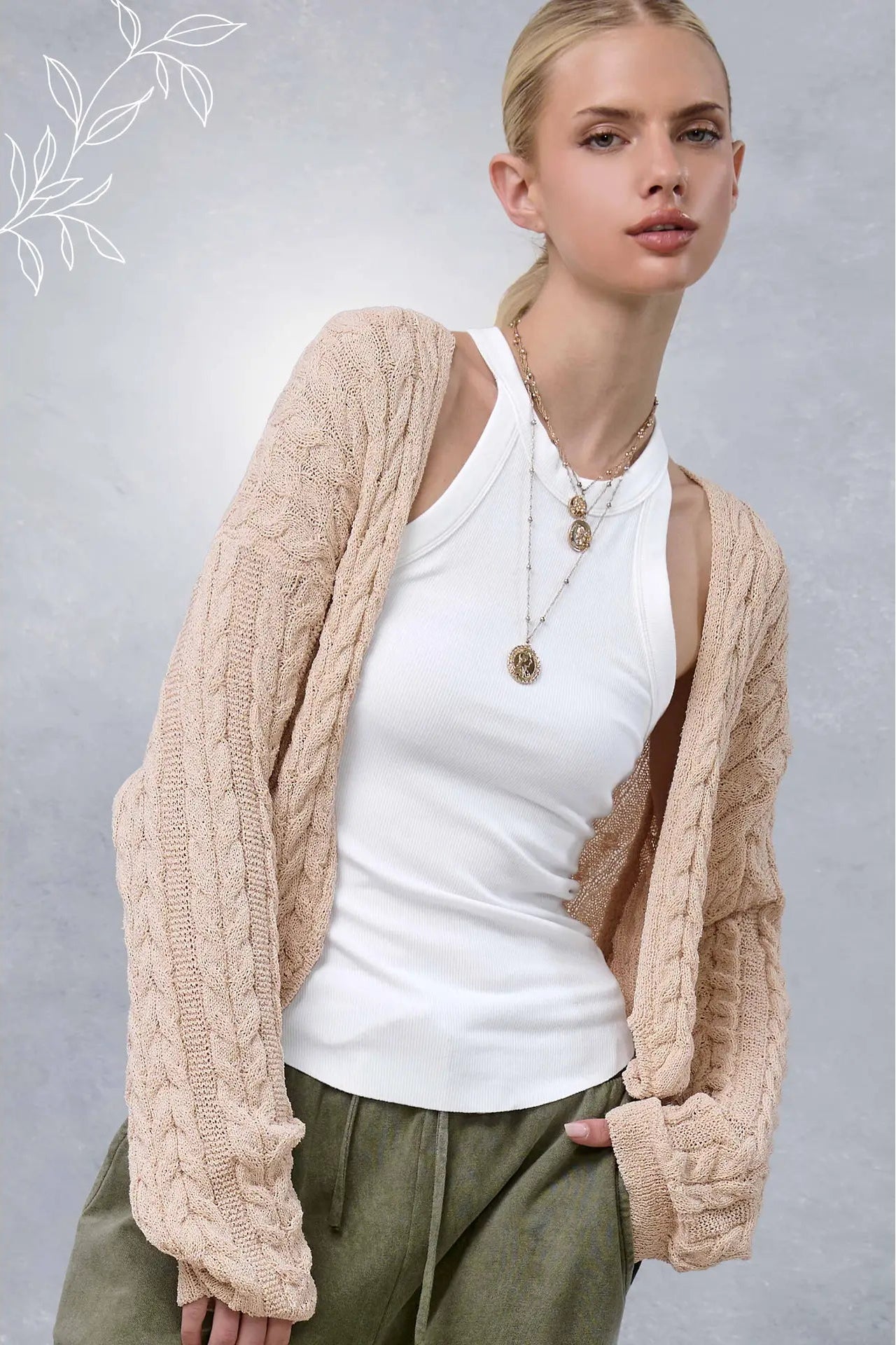 A blonde model wearing a beige cable-knit cardigan over a white tank top and olive green pants, posed against a textured gray background.
