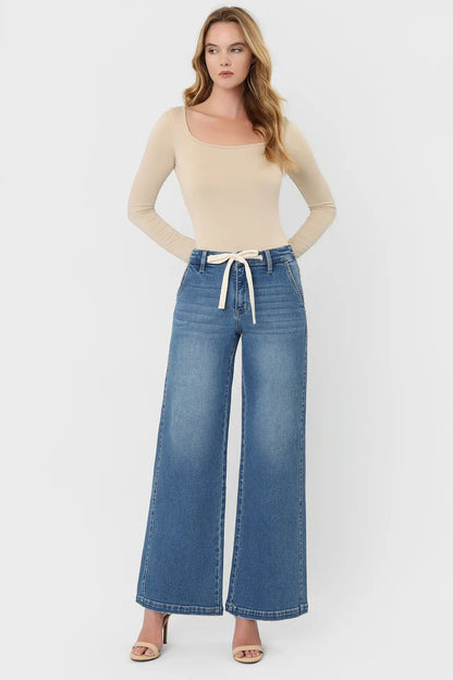 A woman modeling a beige long-sleeve scoop-neck top and high-waisted, wide-leg blue denim jeans with a cream drawstring waist, paired with beige heeled sandals, against a light gray studio background.
