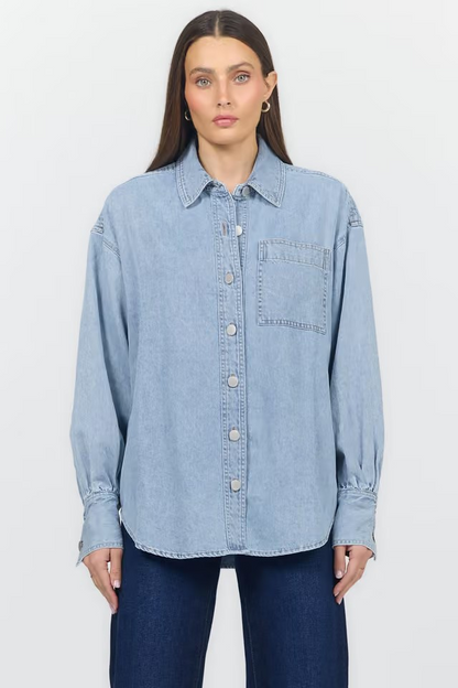 A woman modeling a light blue denim button-up shirt with a chest pocket, paired with dark blue jeans, against a plain light gray studio background.
