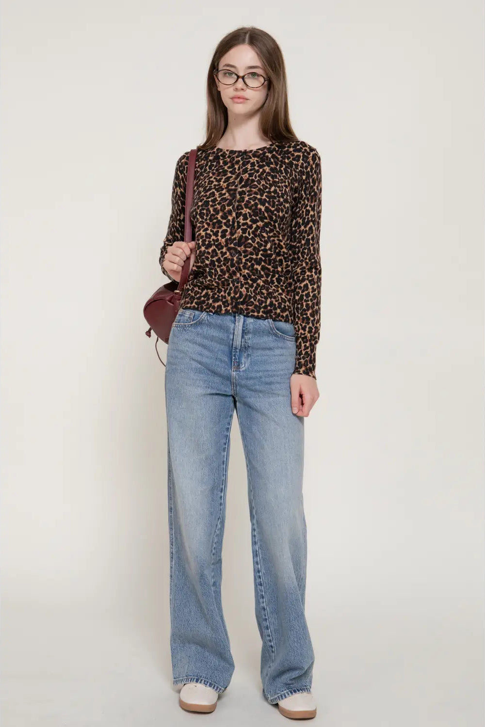 A woman modeling a long-sleeve leopard print top with light-wash wide-leg jeans, glasses, and a small burgundy shoulder bag, standing against a plain light gray studio background.