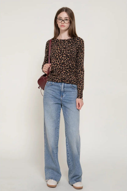 A woman modeling a long-sleeve leopard print top with light-wash wide-leg jeans, glasses, and a small burgundy shoulder bag, standing against a plain light gray studio background.