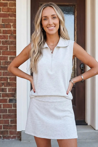 A woman modeling a light heather gray sleeveless polo shirt and matching mini skirt set, standing in front of a brick wall and a dark wooden door.