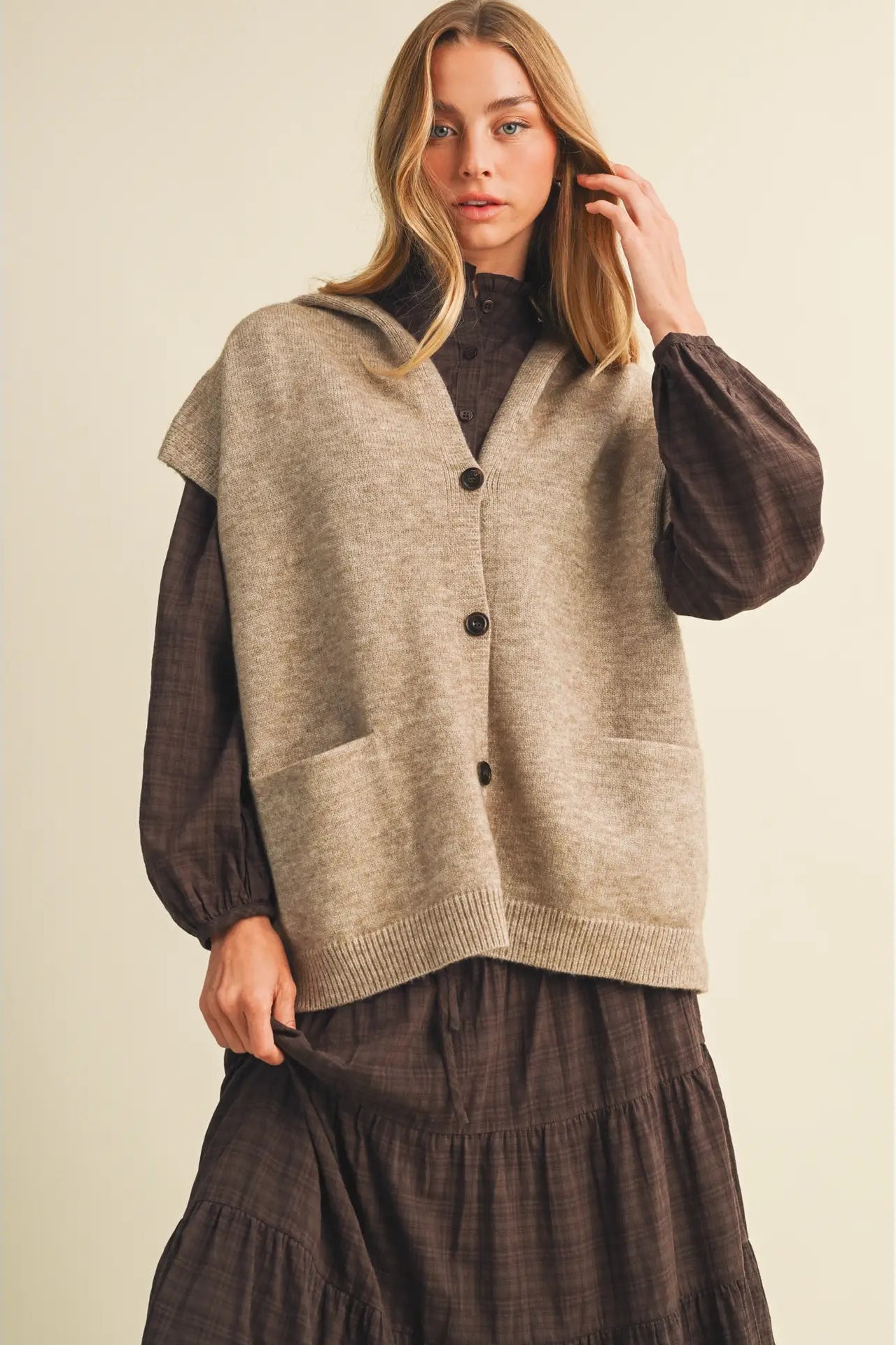 A woman models a sleeveless beige knit cardigan vest with dark buttons over a dark brown long-sleeve top and a matching brown skirt, set against a plain beige background.