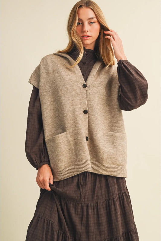 A woman models a sleeveless beige knit cardigan vest with dark buttons over a dark brown long-sleeve top and a matching brown skirt, set against a plain beige background.