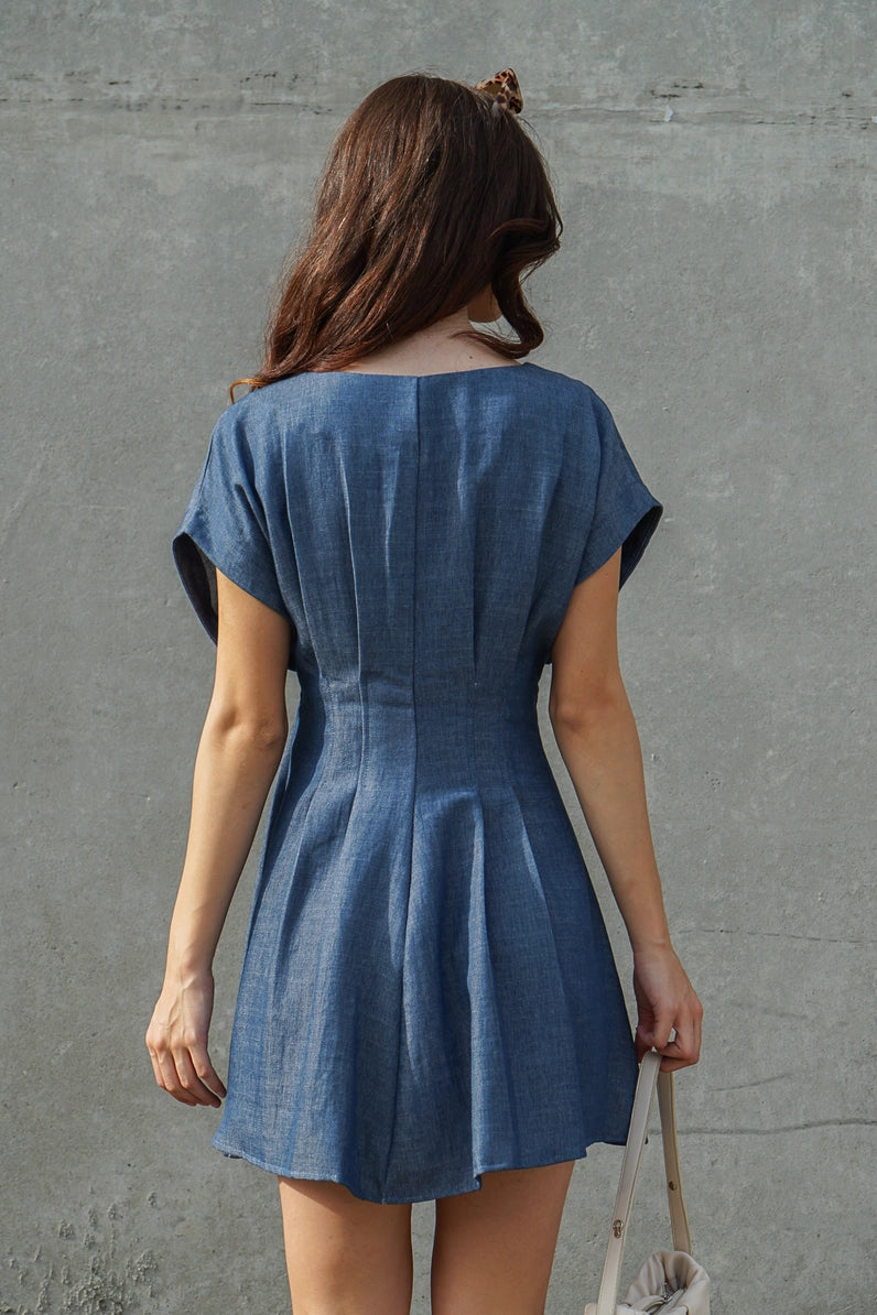 A woman viewed from behind wearing a short-sleeve blue denim-like mini dress with a gathered waist seam, standing against a textured gray concrete wall and holding a light-colored shoulder bag.