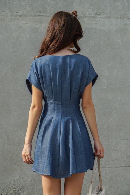 A woman viewed from behind wearing a short-sleeve blue denim-like mini dress with a gathered waist seam, standing against a textured gray concrete wall and holding a light-colored shoulder bag.