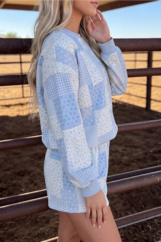 A woman wearing a light blue and white patchwork sweatshirt and matching shorts set, standing in a dirt paddock with metal fences in the background.