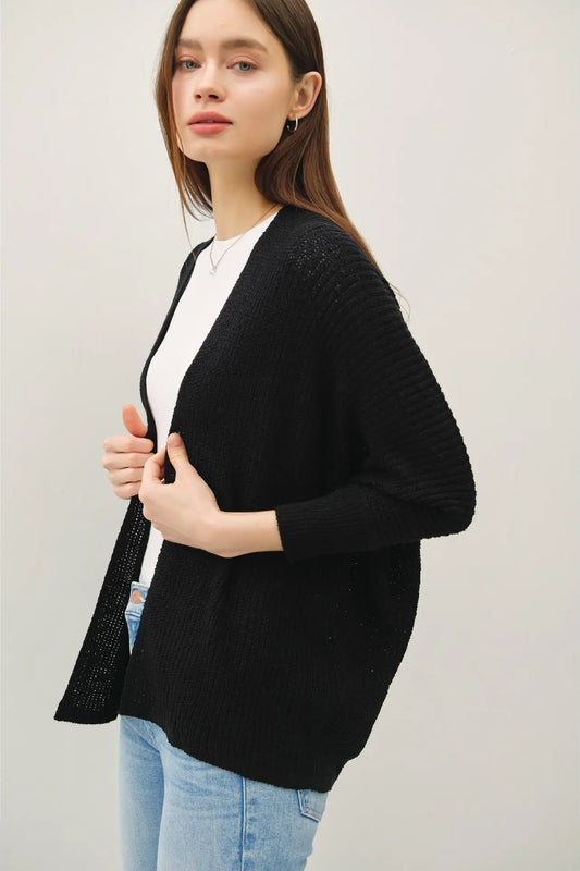 A woman wearing a black open-front knit cardigan over a white top and light blue jeans, photographed against a plain light gray studio background.