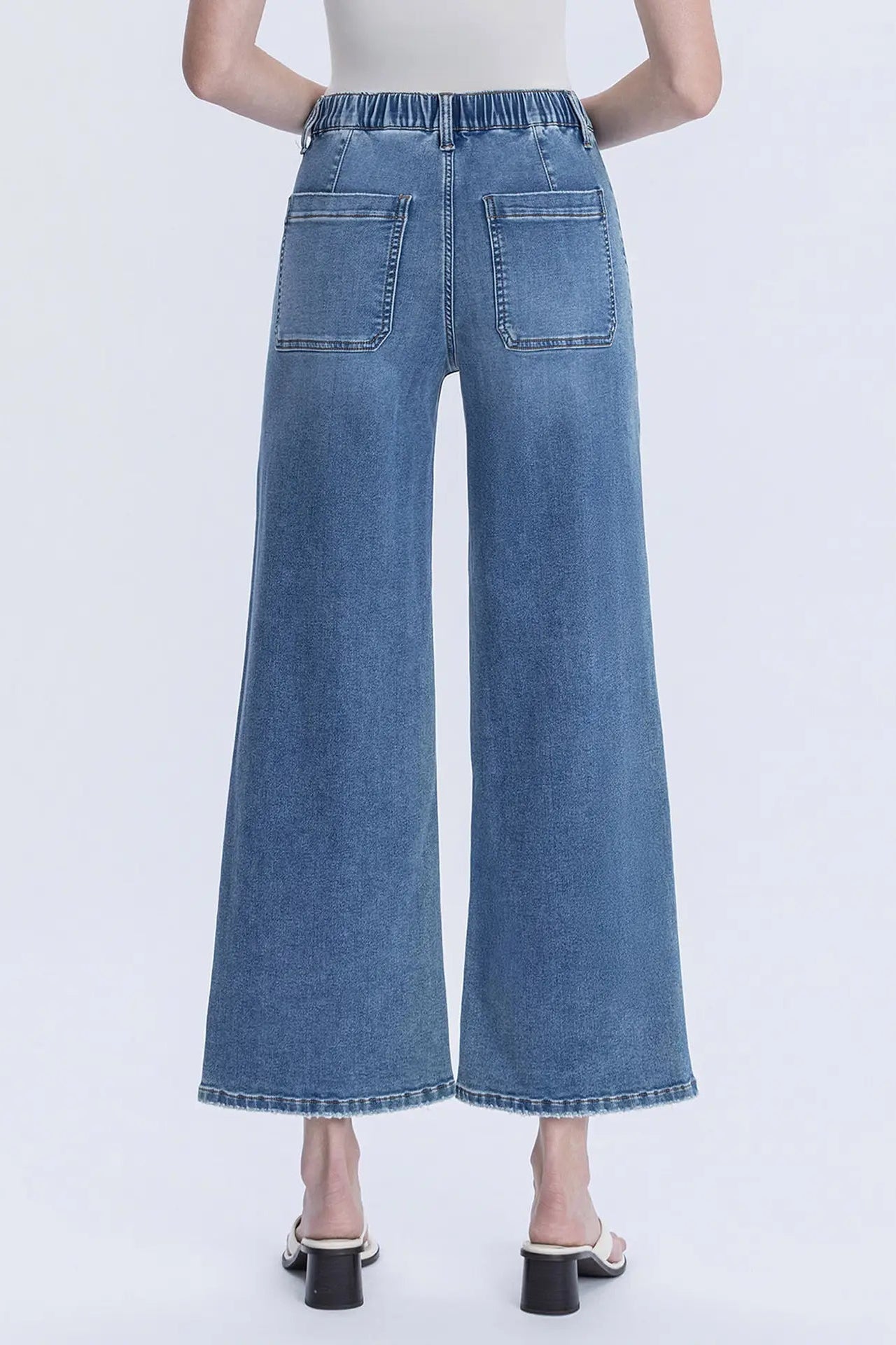 Back view of a model wearing high-waisted, wide-leg blue denim jeans with a button and zip fly, rear patch pockets, and a relaxed straight-leg fit, paired with black heeled sandals against a light gray studio background.