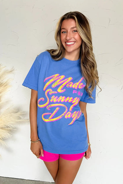 Woman wearing a blue t-shirt with 'Made for Sunny Days' text, standing against a white wall.