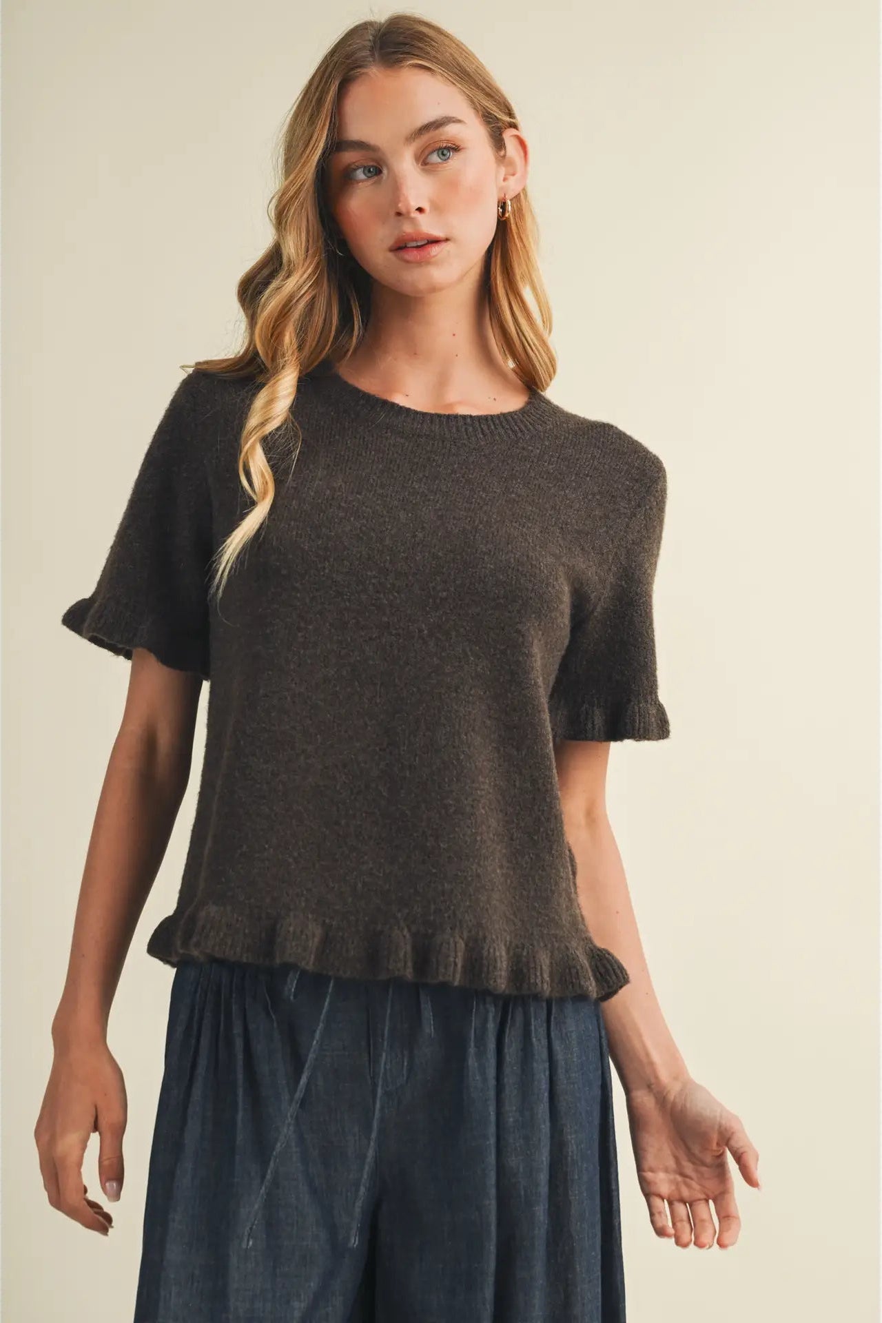 A woman modeling a dark brown short-sleeve knit top with ruffled cuffs and hem, paired with dark blue wide-leg pants, against a solid beige background.