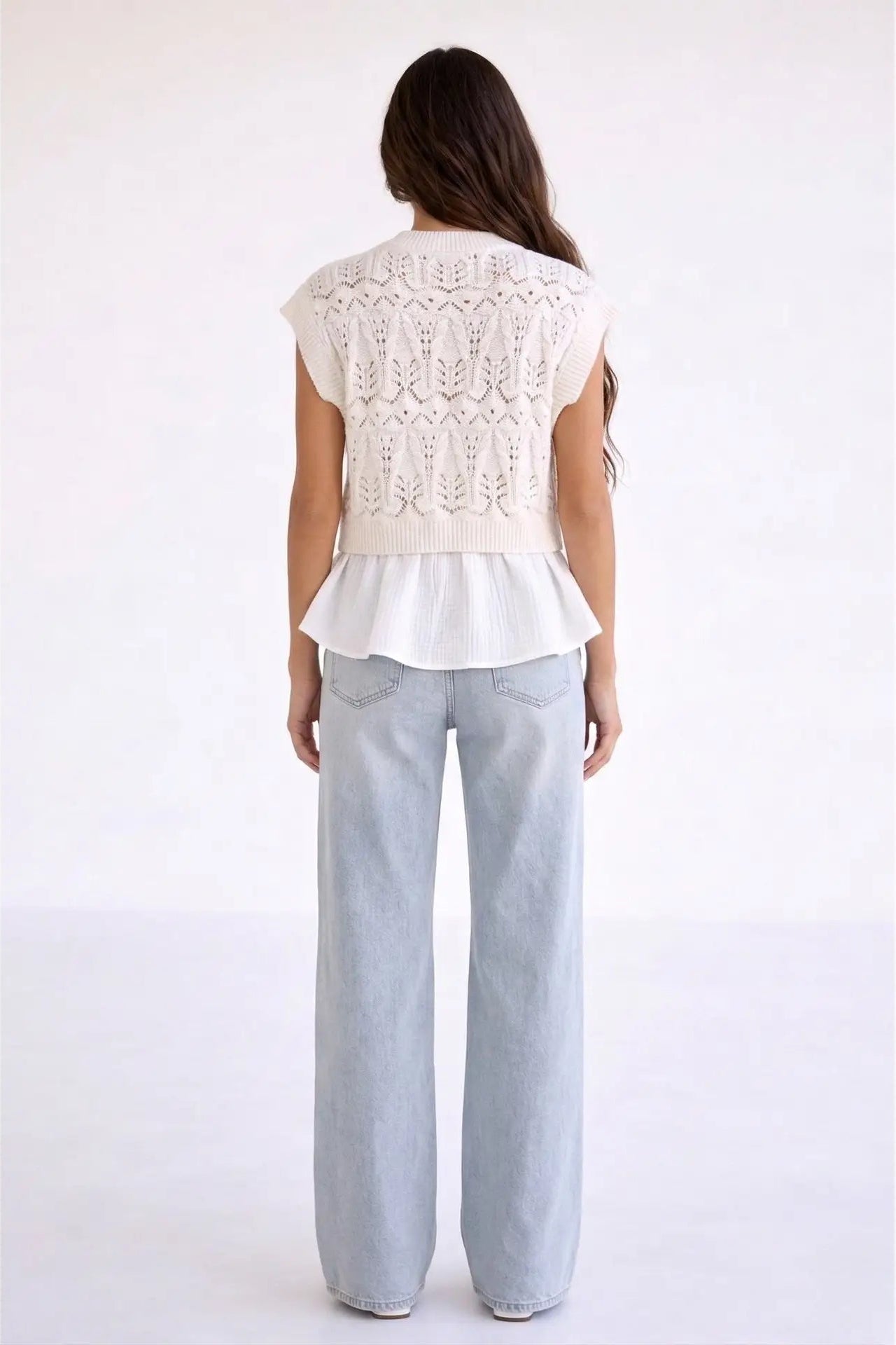 A woman viewed from the back wearing a white sleeveless knit top with a textured pattern and ruffled hem, paired with light-wash wide-leg jeans, against a plain white studio background.