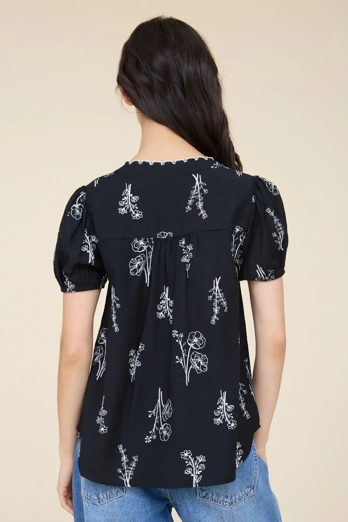 A woman viewed from the back wearing a black short-sleeve blouse with a white floral print and puffed sleeves, paired with blue jeans against a plain beige background.