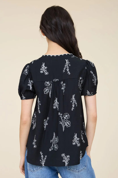 A woman viewed from the back wearing a black short-sleeve blouse with a white floral print and puffed sleeves, paired with blue jeans against a plain beige background.