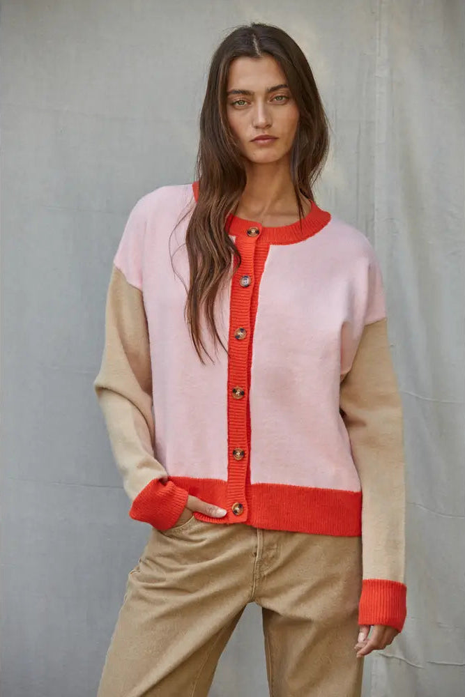 A woman wearing a colorblock cardigan sweater with a light pink body, beige sleeves and lower hem, and a bright red-orange ribbed trim and button placket, paired with beige pants against a gray backdrop.