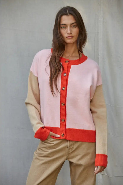 A woman wearing a colorblock cardigan sweater with a light pink body, beige sleeves and lower hem, and a bright red-orange ribbed trim and button placket, paired with beige pants against a gray backdrop.