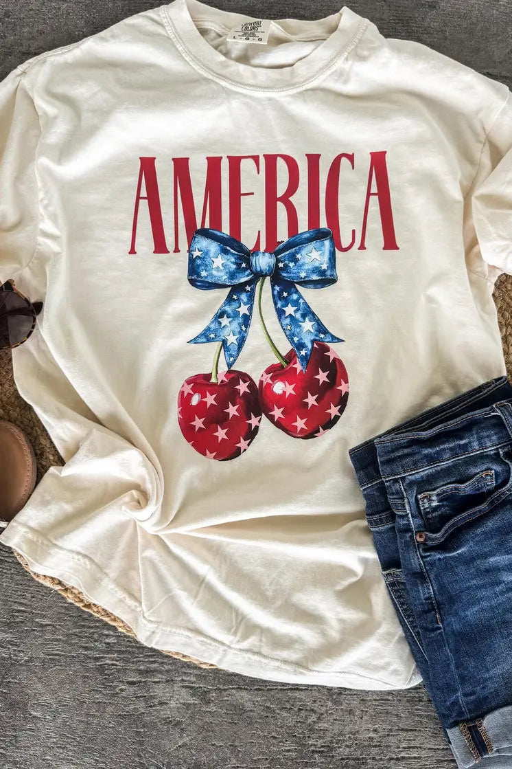 White t-shirt with 'AMERICA' text and cherry design on a wooden surface
