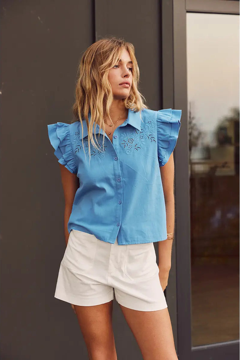 Woman wearing a blue blouse with ruffled sleeves and white shorts standing against a neutral background.