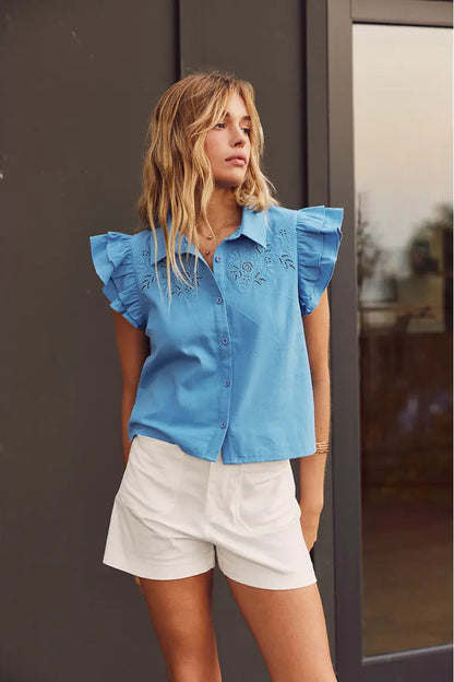 Woman wearing a blue blouse with ruffled sleeves and white shorts standing against a neutral background.