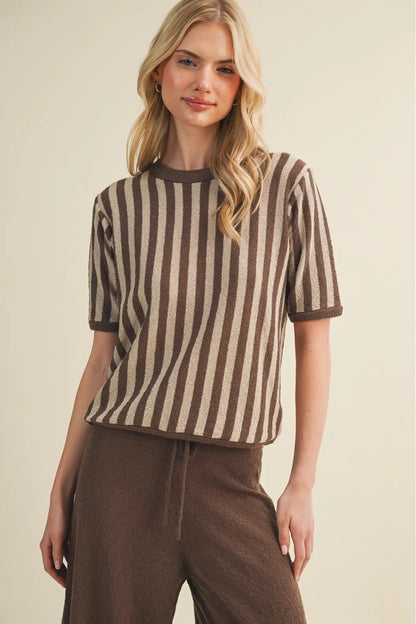 A woman modeling a short-sleeve knit top with vertical brown and beige stripes, paired with brown drawstring pants, against a plain beige studio background.