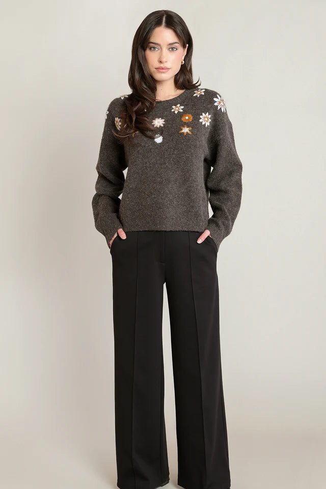 A woman modeling a dark gray knit sweater with embroidered floral details on the chest, paired with black wide-leg trousers, against a neutral light gray background.