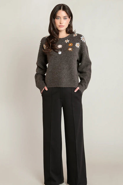 A woman modeling a dark gray knit sweater with embroidered floral details on the chest, paired with black wide-leg trousers, against a neutral light gray background.