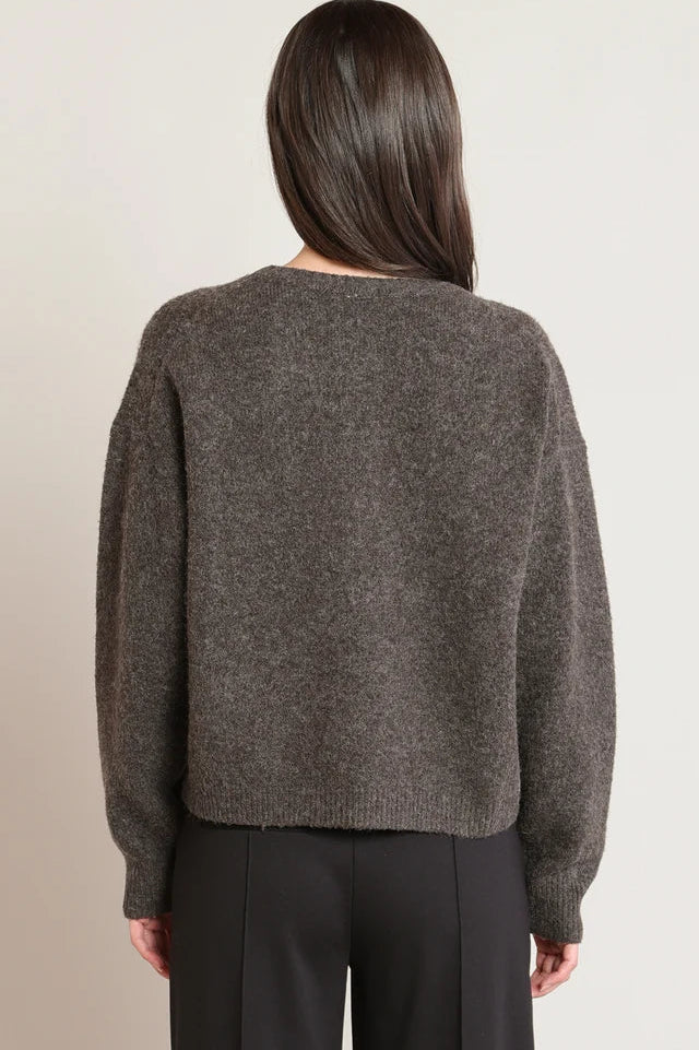 A woman viewed from the back wearing a dark brown textured knit sweater and black trousers, set against a plain light beige background.