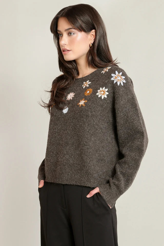 A woman wearing a dark brown textured knit sweater with embroidered daisy and flower motifs on the chest, paired with black trousers, set against a plain light gray studio background.