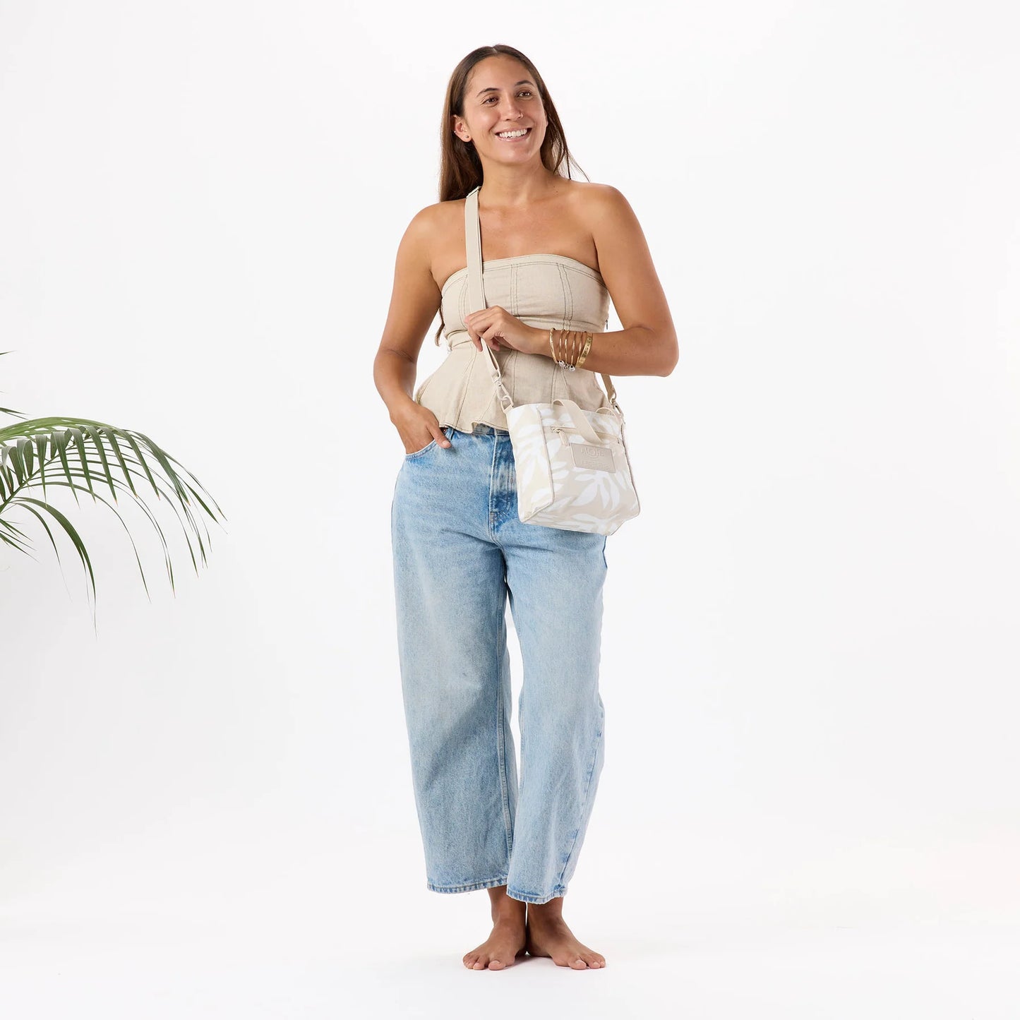 Woman wearing a beige strapless top and light blue jeans, holding a white bag on a white background.