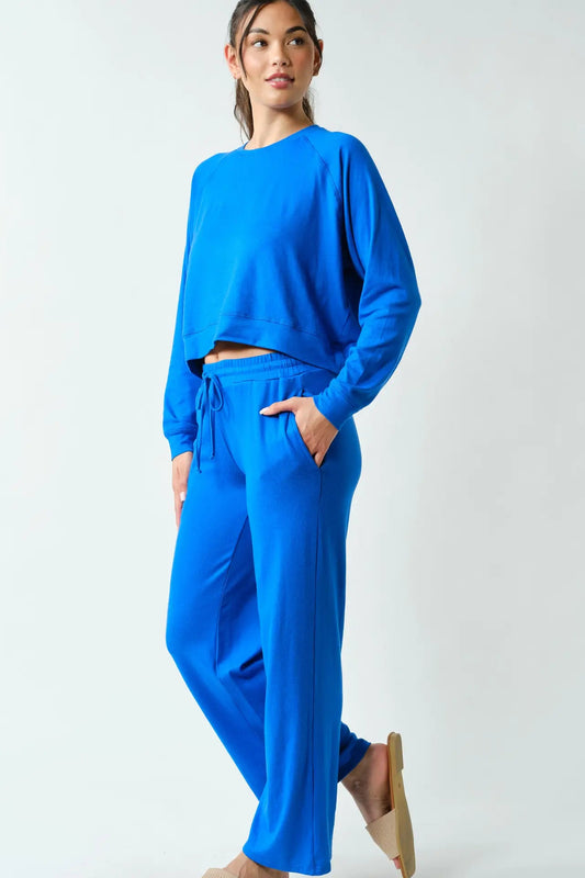A woman modeling a bright blue two-piece loungewear set consisting of a cropped long-sleeve top and matching drawstring pants, standing against a plain light gray studio background.