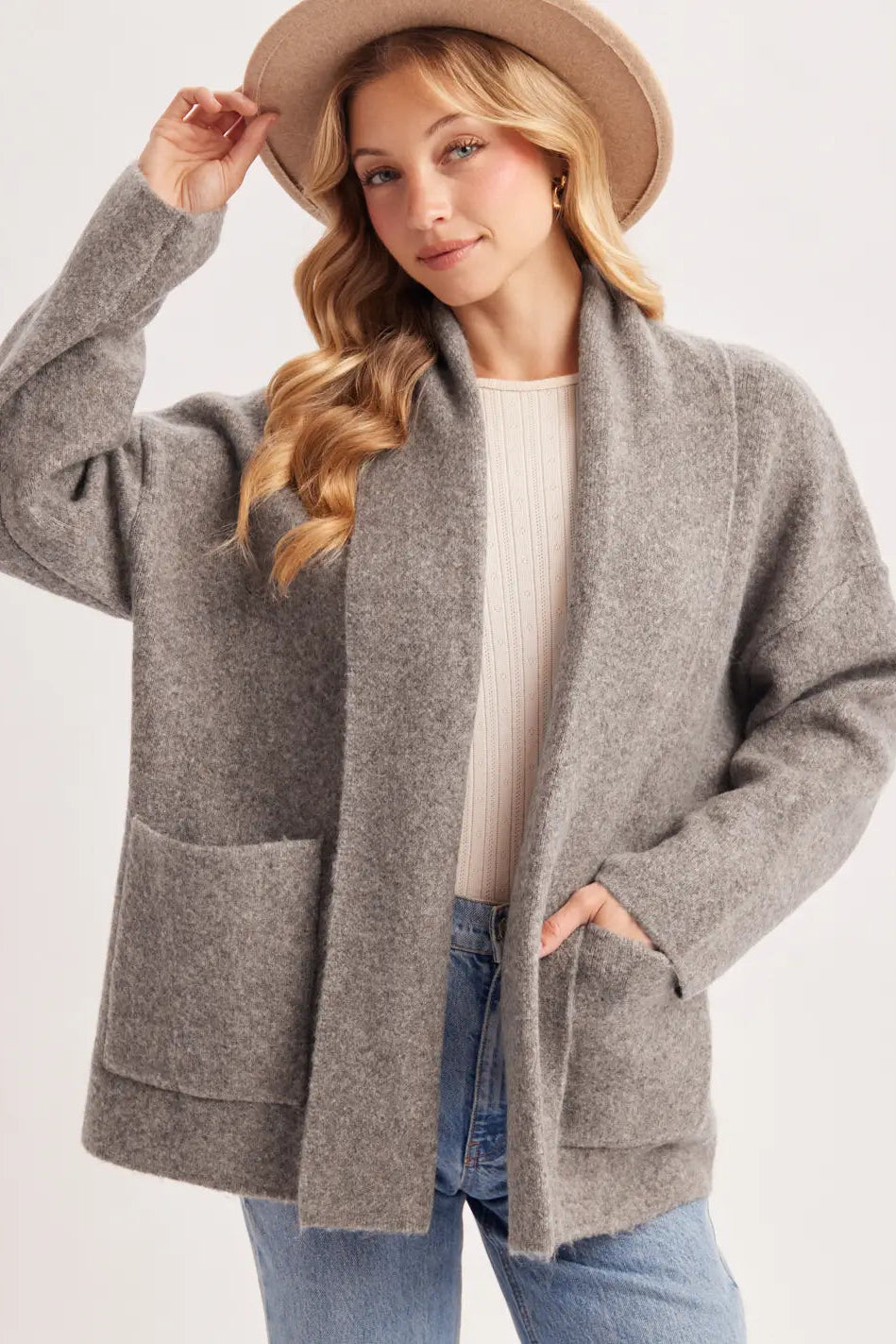 A woman wearing a heather gray open-front cardigan with large front pockets, a cream ribbed top, blue jeans, and a tan wide-brim hat, photographed against a light gray studio background.